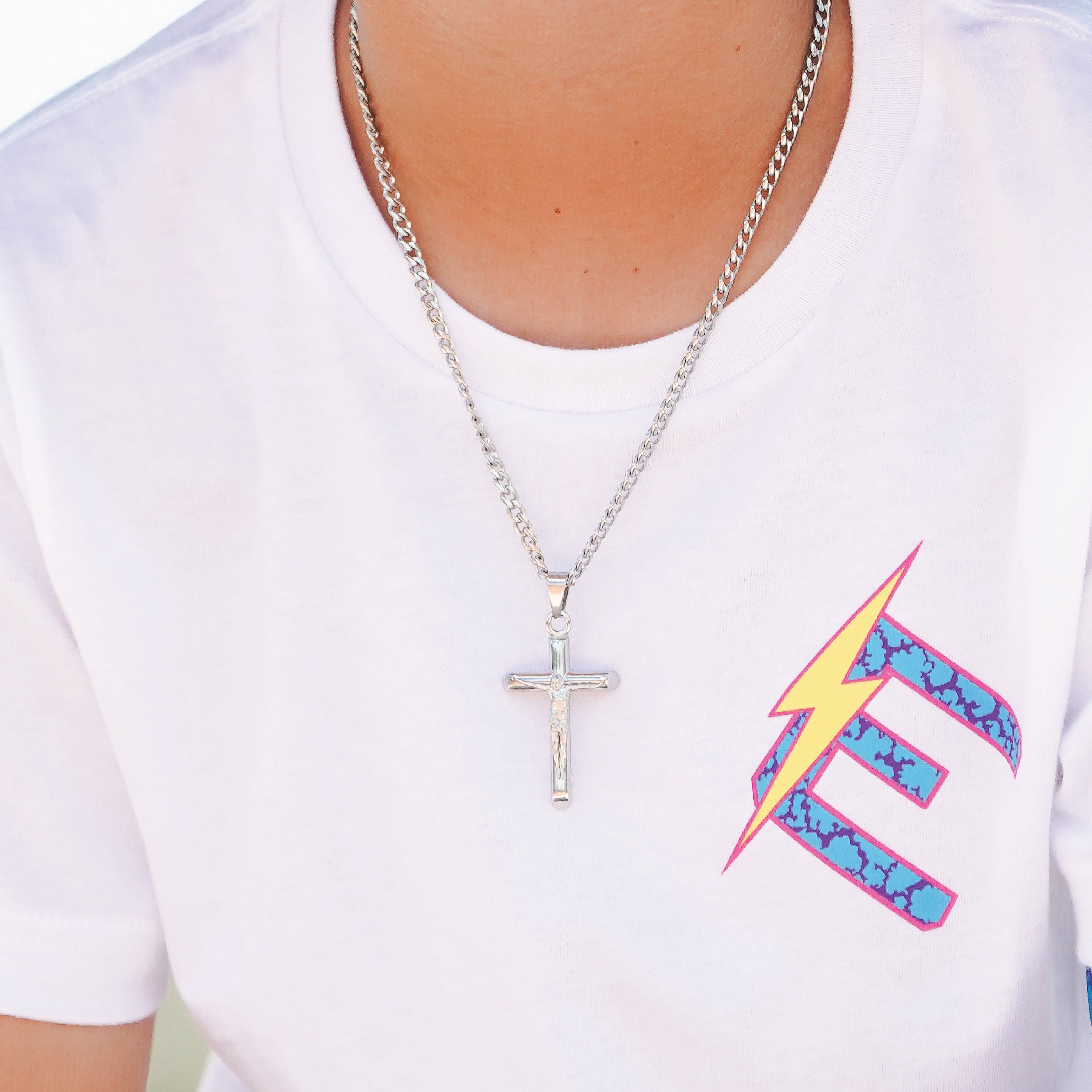A silver crucifix pendant necklace worn by a person in a white t-shirt featuring a colorful graphic print.