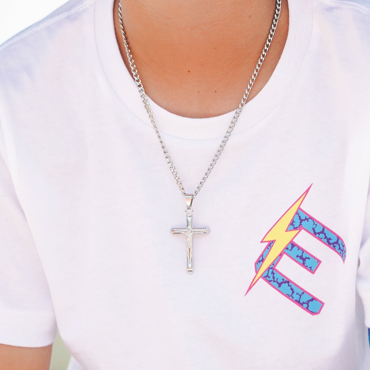 A silver crucifix pendant necklace worn by a person in a white t-shirt featuring a colorful graphic print.