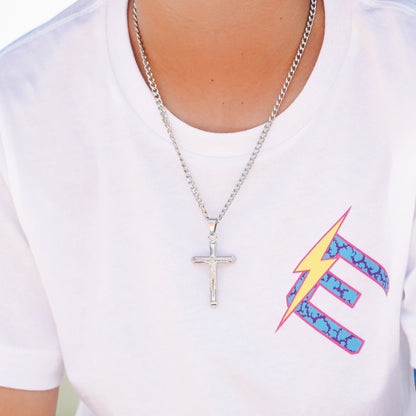 A silver crucifix pendant necklace worn by a person in a white t-shirt featuring a colorful graphic print.